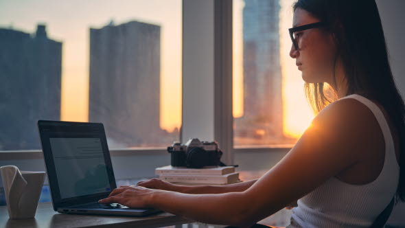 Young woman working at her computer with view of skyscrapers outside the window