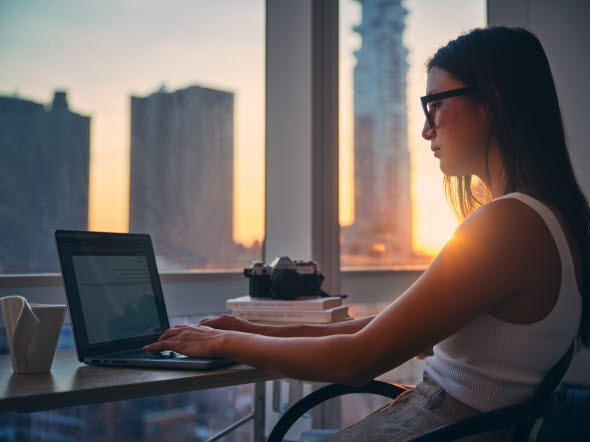 Young woman working at her computer with view of skyscrapers outside the window