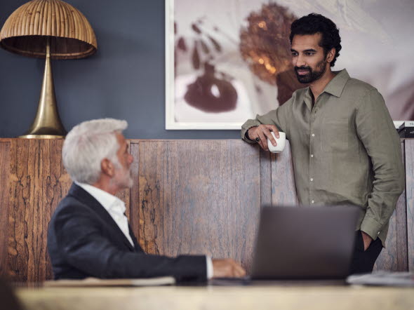 Picture of two men talking to each other, one seated on a sofa in front of a computer