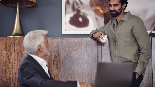 Picture of two men talking to each other, one seated on a sofa in front of a computer